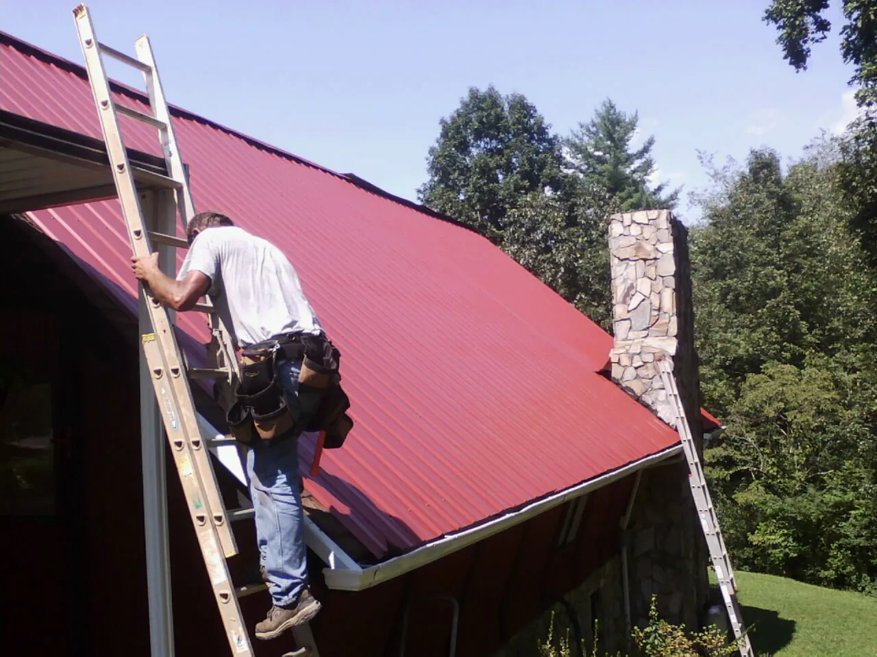 Roofer on a ladder beside a red metal roof and stone chimney — Soffit & Fascia Repair by Roofing Brothers in Constableville